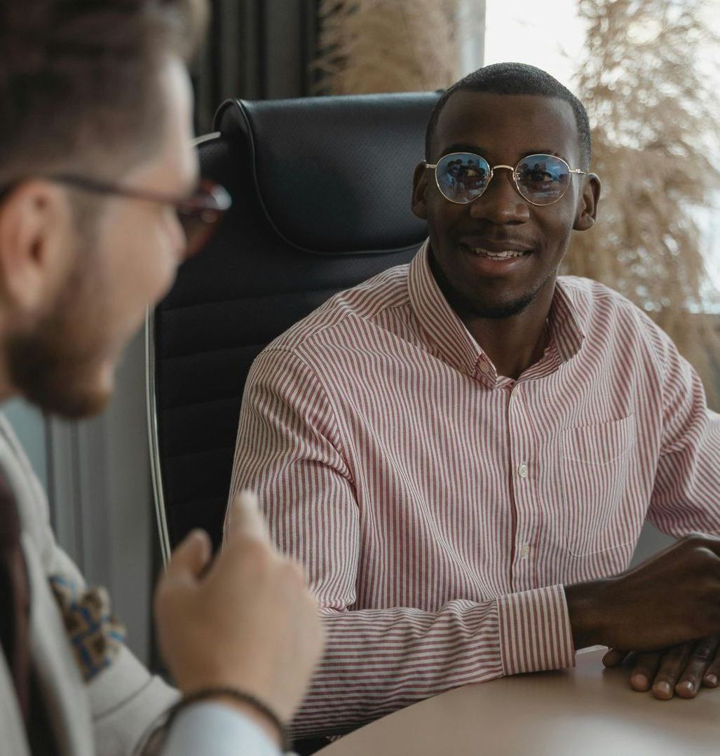 A man wearing glasses is sitting at a table talking to another man.