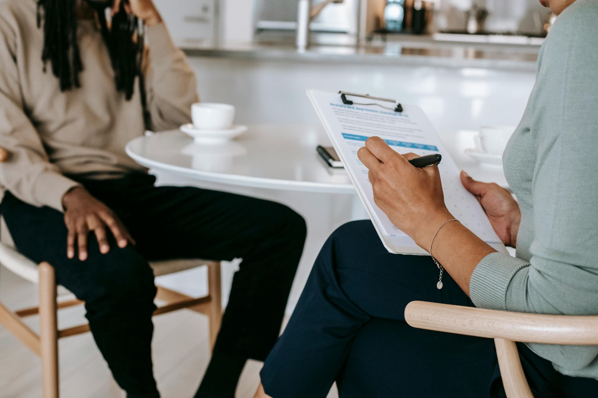 A woman is sitting at a table talking to another woman while holding a clipboard.