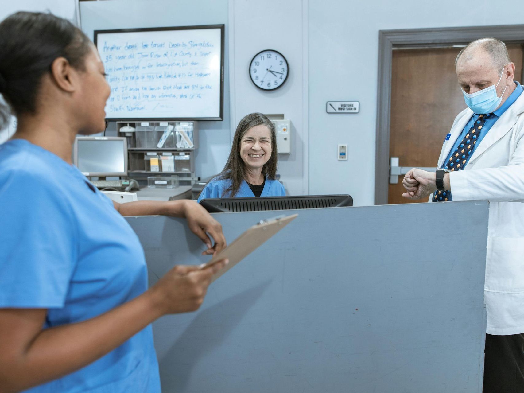 A doctor wearing a mask is talking to a nurse at a hospital reception desk.