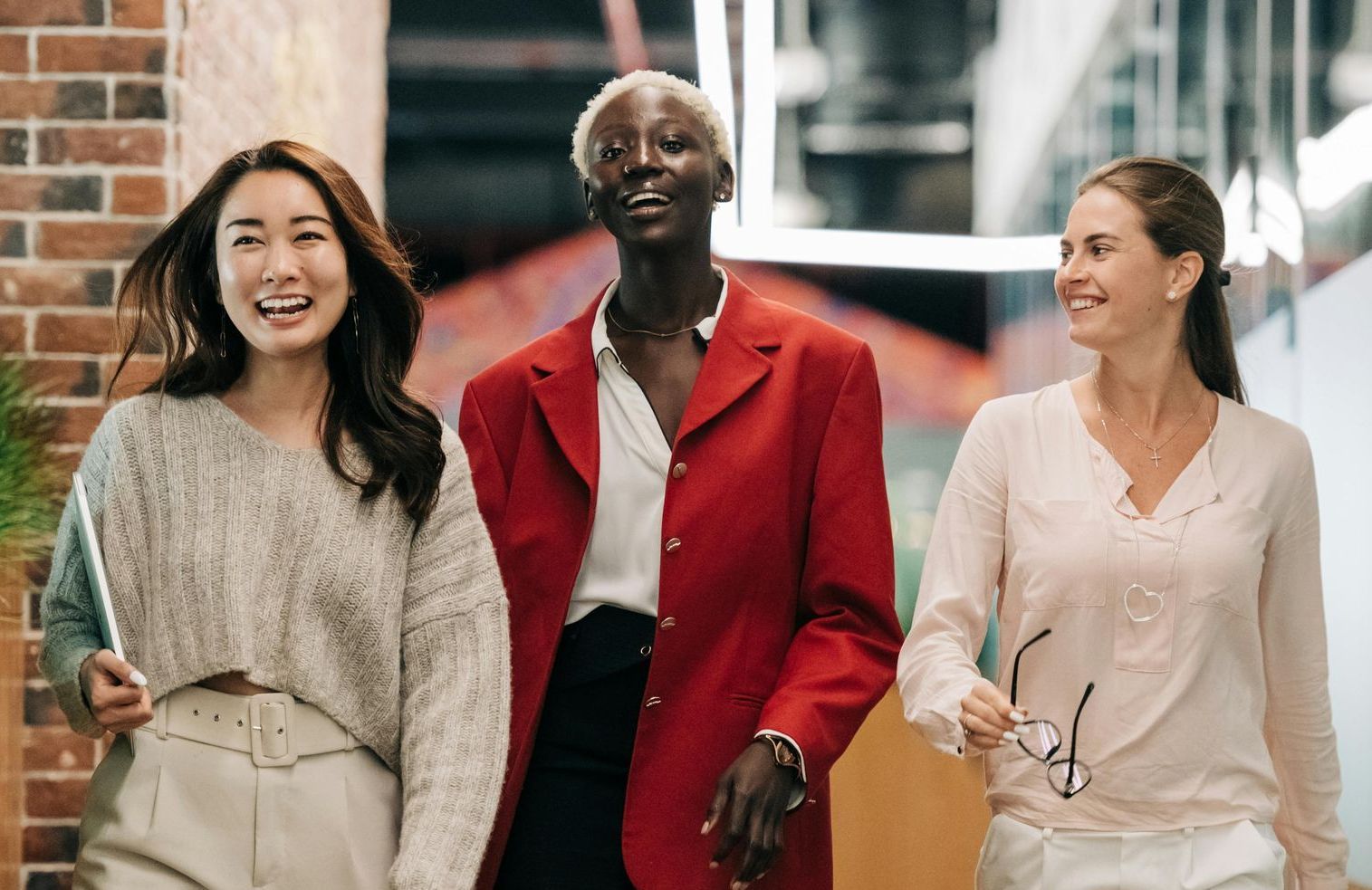 Three women are walking down a hallway holding hands and smiling.