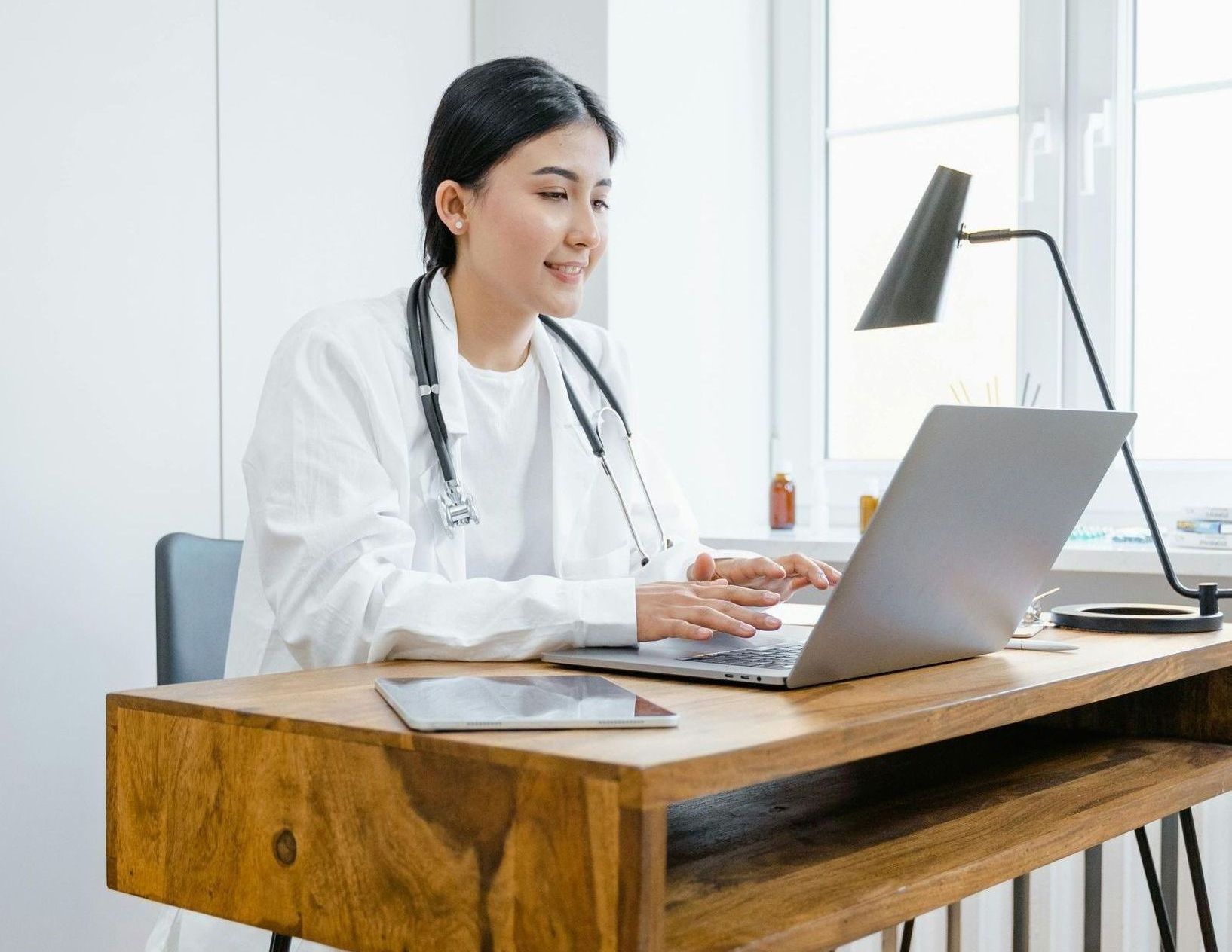 A female doctor is sitting at a desk using a laptop computer.