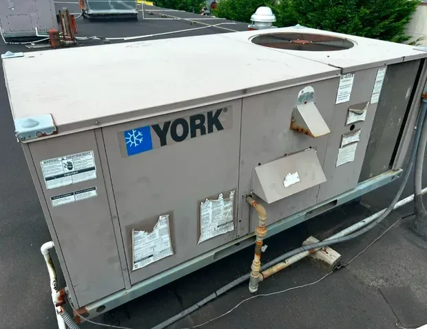 A weathered York rooftop HVAC unit with metallic casing, sitting on a flat roof next to piping and electrical conduits.