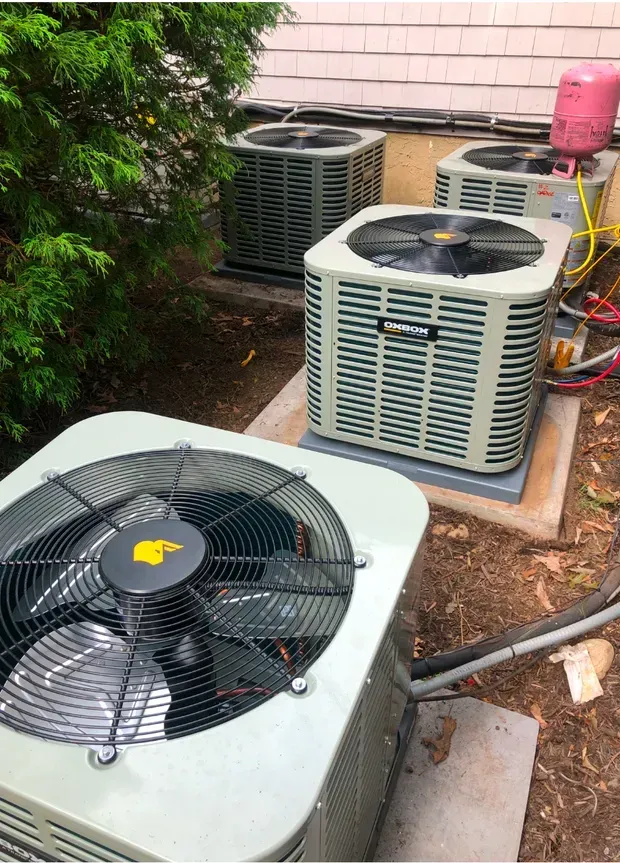 Four outdoor HVAC condenser units sit on concrete pads next to a building and evergreen hedge.