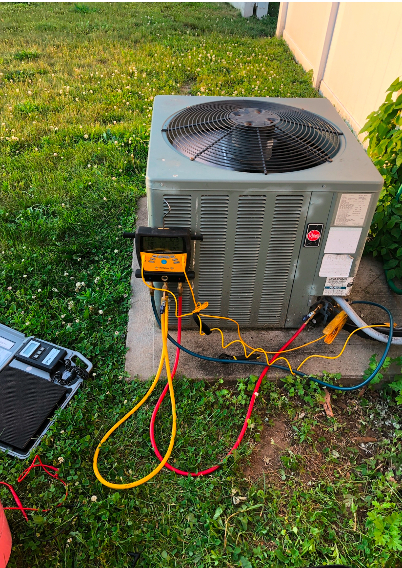 An air conditioning condenser unit sits on a concrete pad in a grassy yard, connected to yellow and red diagnostic gauges.
