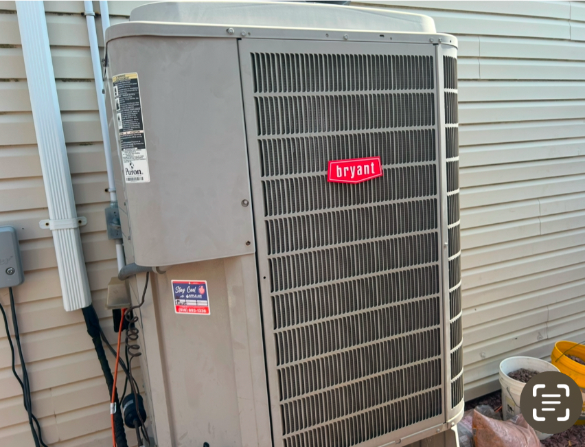 A beige Bryant HVAC outdoor condenser unit sitting against a light-colored vinyl-sided wall.