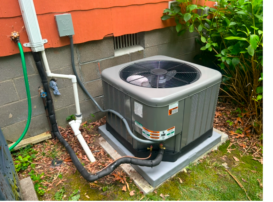 A gray residential HVAC outdoor unit sitting on a concrete pad next to an orange exterior wall with plumbing and foliage.