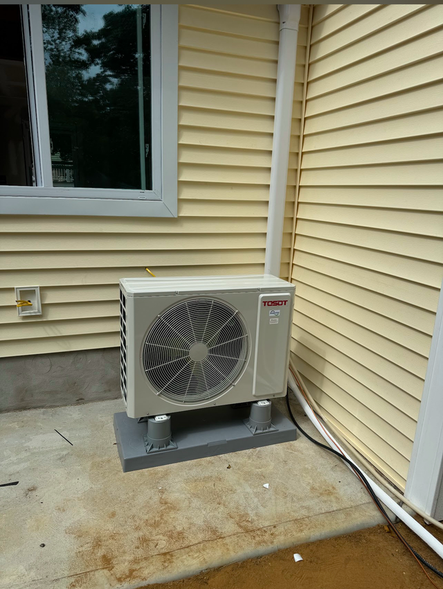 A light-colored outdoor HVAC condenser unit sits on a concrete pad against a house with yellow vinyl siding.