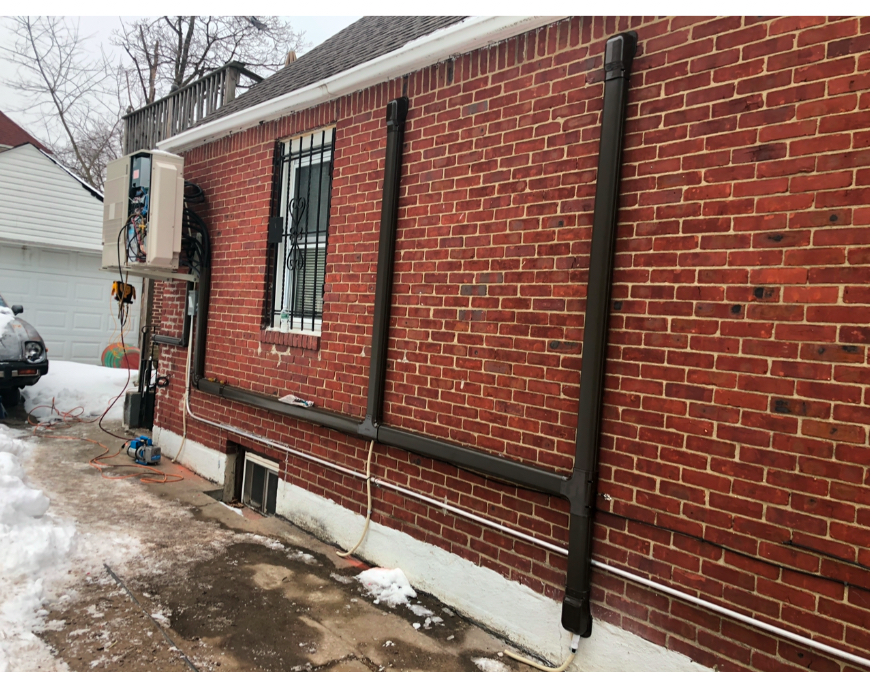 A brick building exterior with a mounted HVAC unit and dark protective conduit running along the wall to ground level.