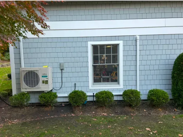 A light-gray shingled wall with a central white-framed window, a Fujitsu HVAC unit, and a row of small, round green bushes.