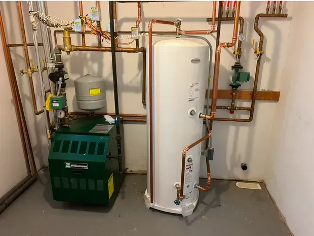 A green boiler stands next to a white hot water tank in a utility room with copper plumbing connections on the wall.