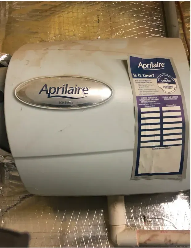 A light-colored Aprilaire humidifier unit mounted on a reflective wall with a service sticker on the right side.