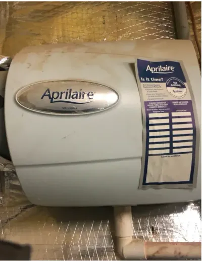 A light-colored Aprilaire humidifier unit mounted on a reflective wall with a service sticker on the right side.
