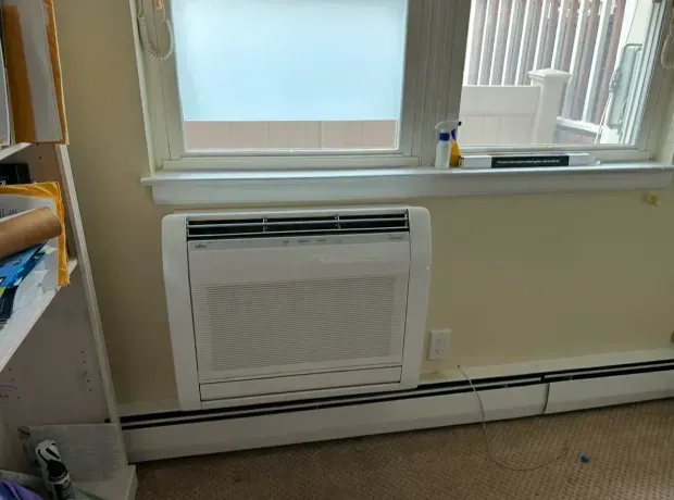 A white floor-mounted air conditioning unit installed beneath a window in a room with beige walls and carpeted flooring.