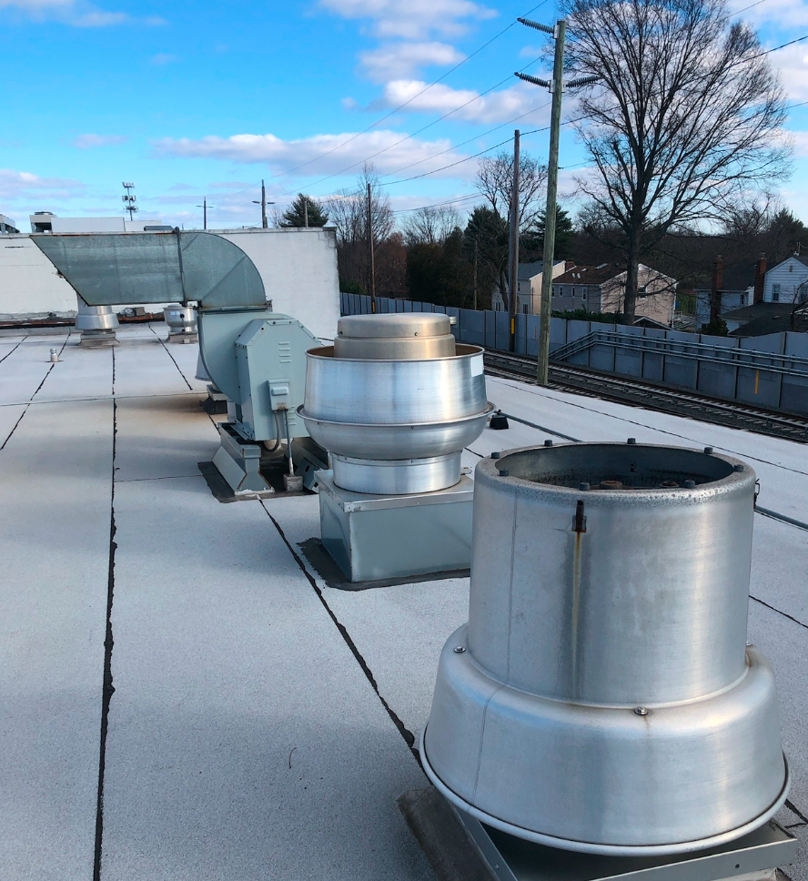 Metallic industrial roof exhaust fans and ductwork on a flat roof against a blue sky with trees in the background.