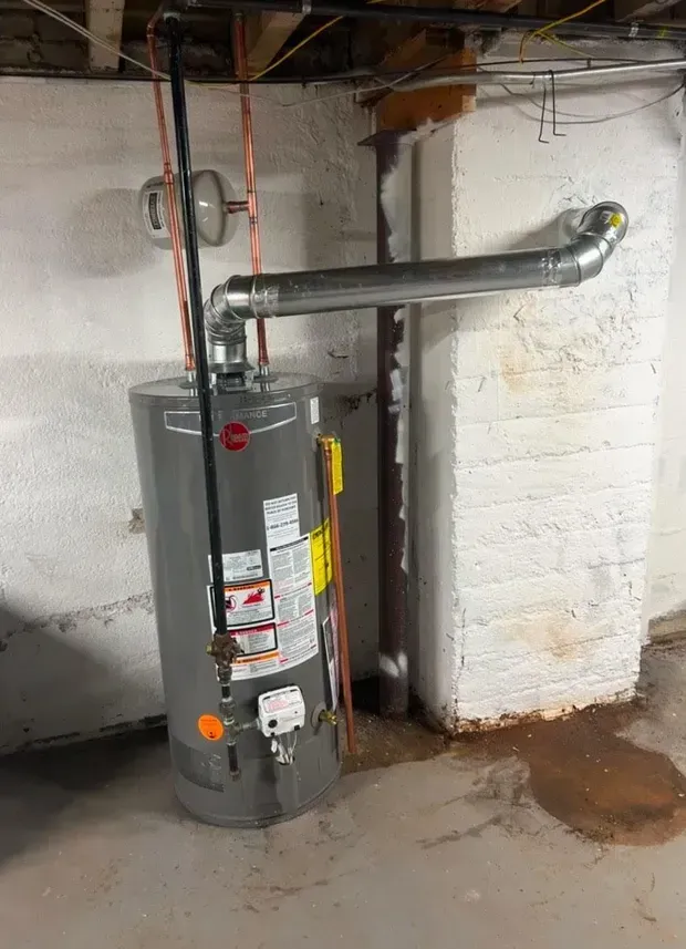 A gray gas water heater stands in a basement against a white wall, connected to a metal vent pipe and copper plumbing.