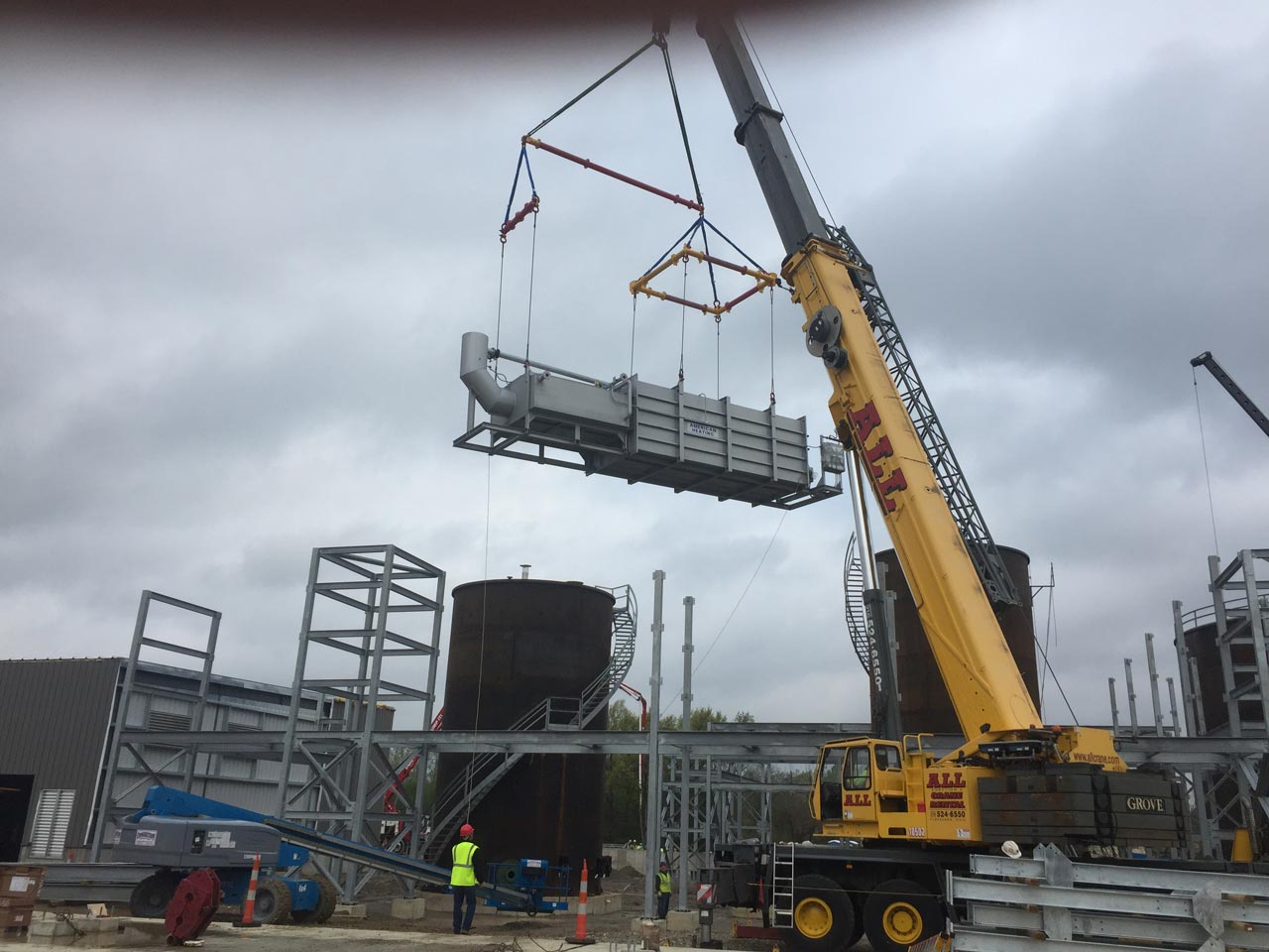 A crane lifts industrial equipment at a construction site with gray skies.