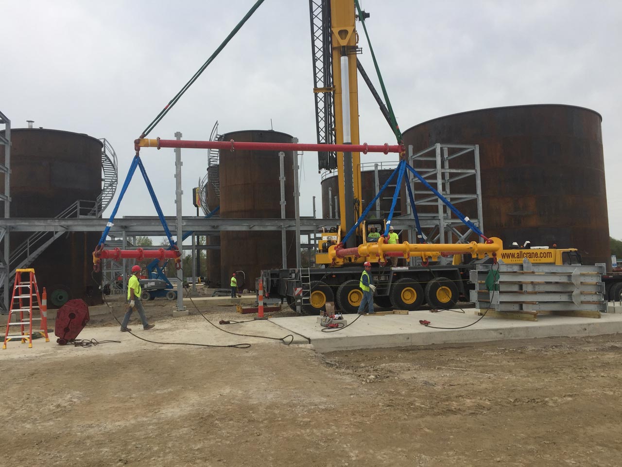 Crane lifting metal structure at industrial site with tanks and workers.