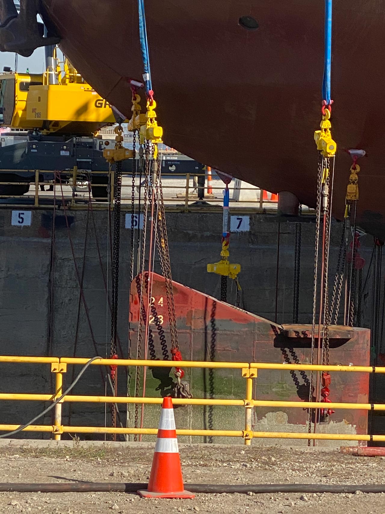 Ship stern being lifted by chains, dockside. Red hull, yellow safety rails, and an orange traffic cone.