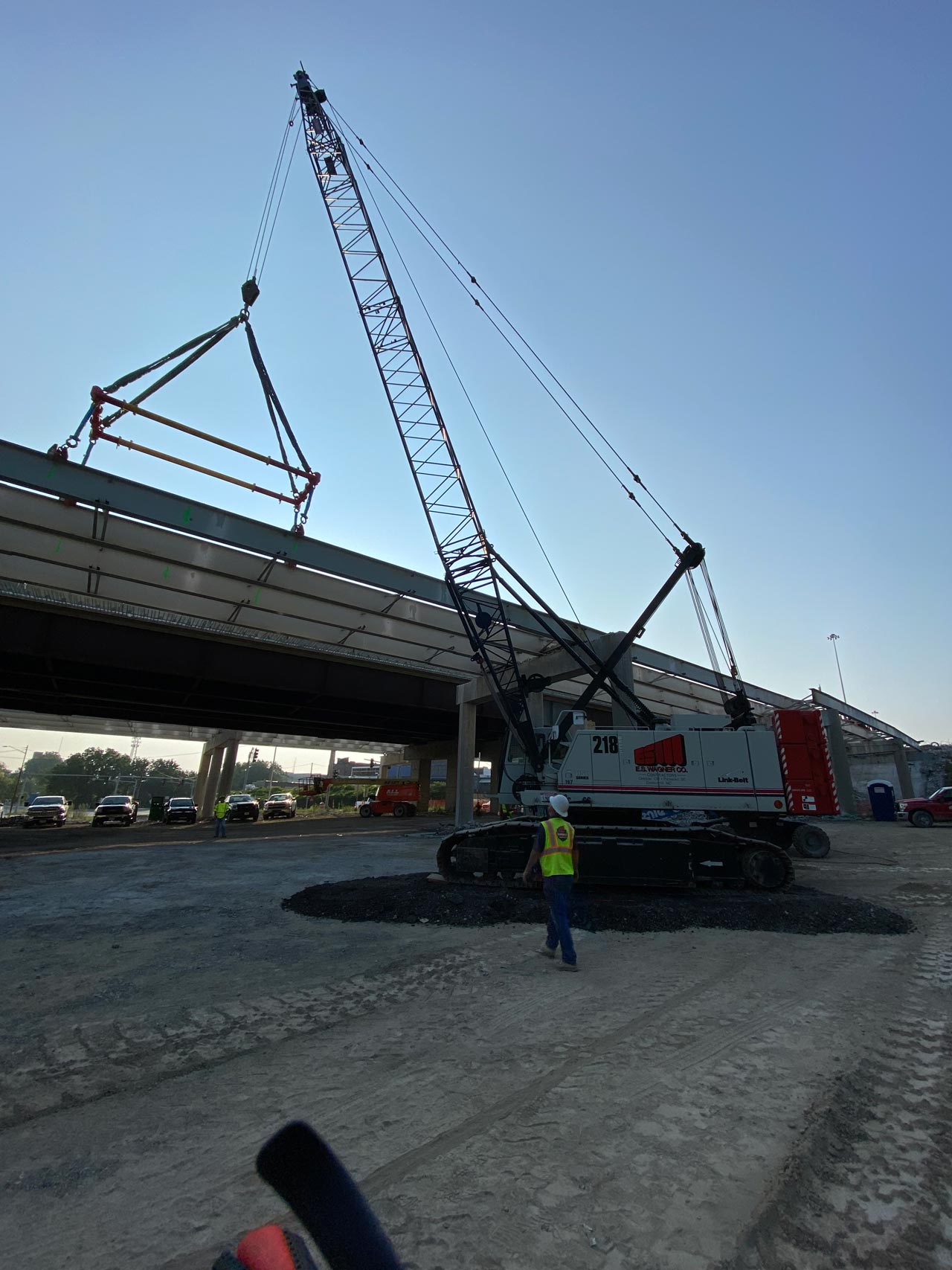 A large crane lifting a bridge beam at a construction site. A worker in a hard hat watches.