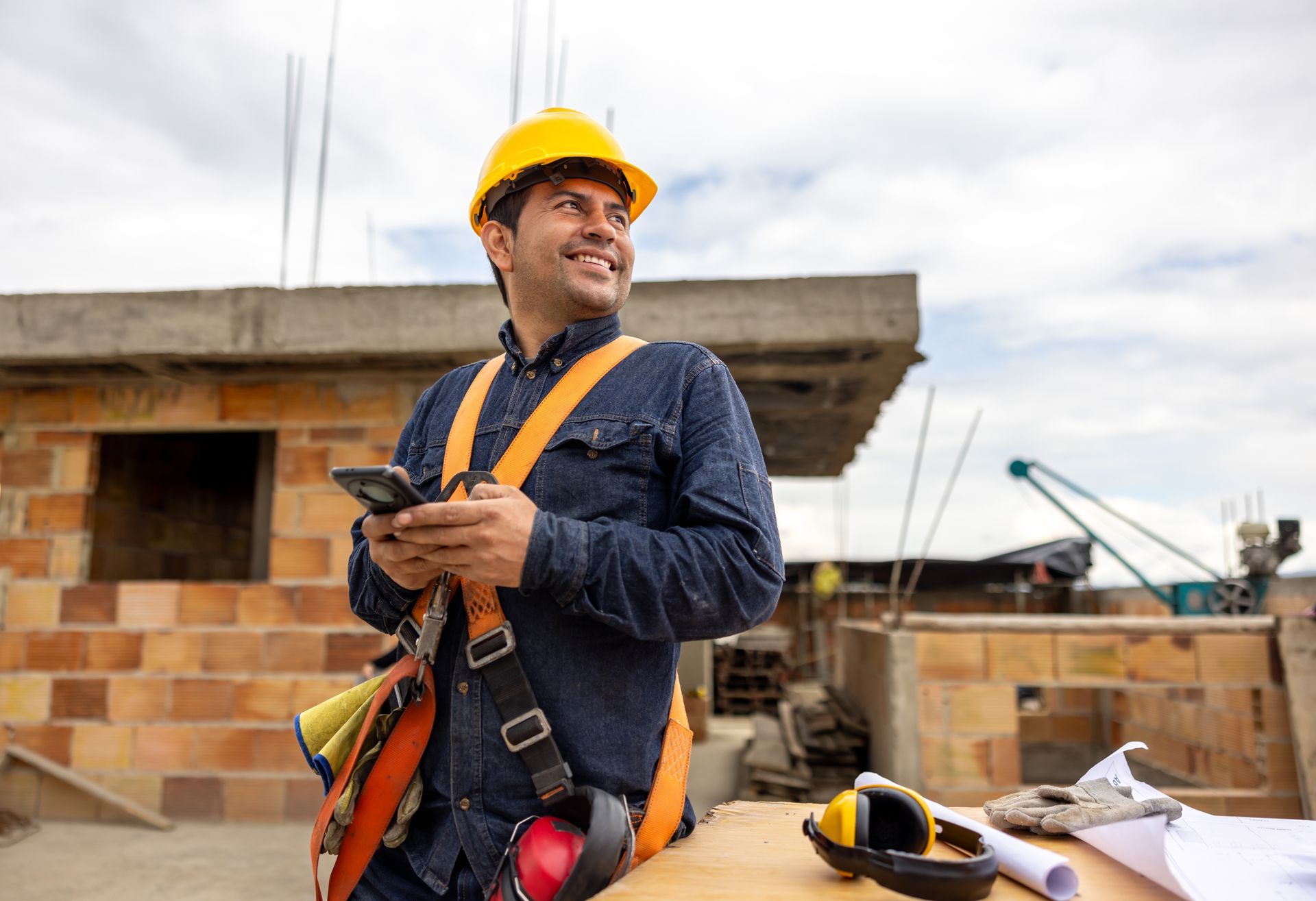 A construction worker, with safety gear on, is smiling and using his cell phone at a building site.