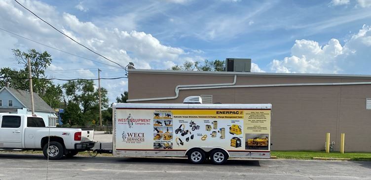 A white truck towing a trailer with tools parked near a beige building under a partly cloudy sky.
