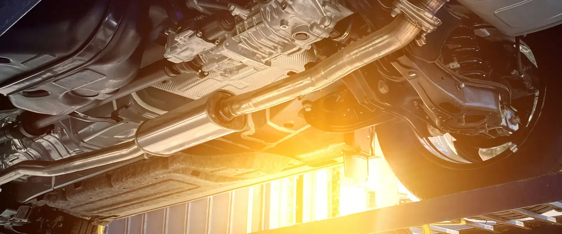 Undercarriage of a vehicle, showing exhaust system, suspension, and tire from below, lit by a bright light.