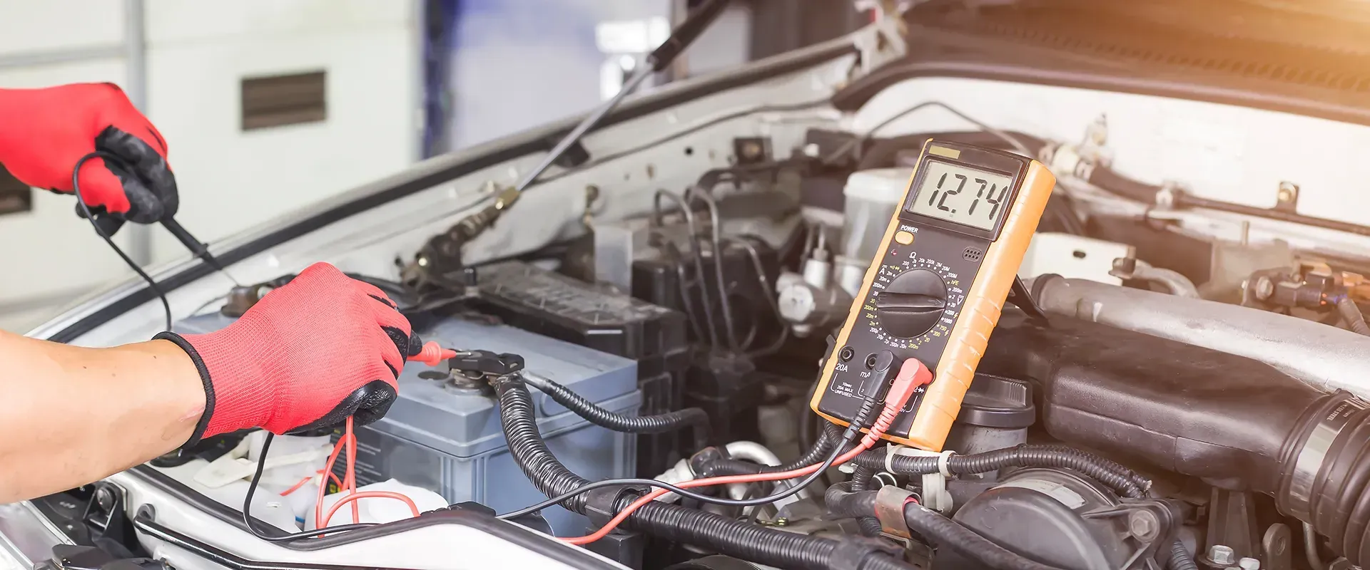 Person wearing red gloves using a multimeter on a car battery to check its voltage.