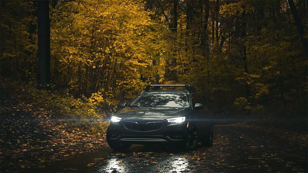 Black SUV driving on a wet forest road, surrounded by autumn foliage; headlights on.