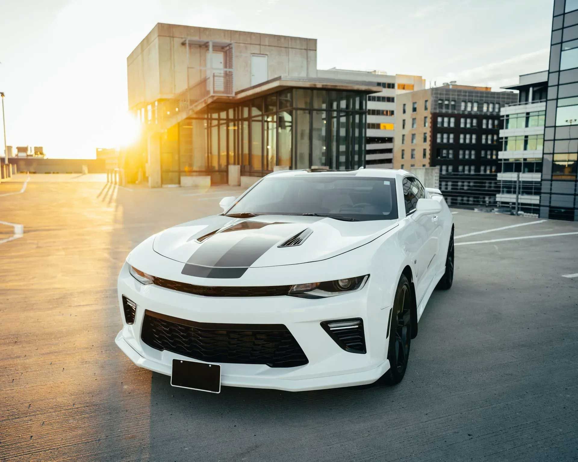 White Chevrolet Camaro with black racing stripes parked on a rooftop parking lot at sunset.