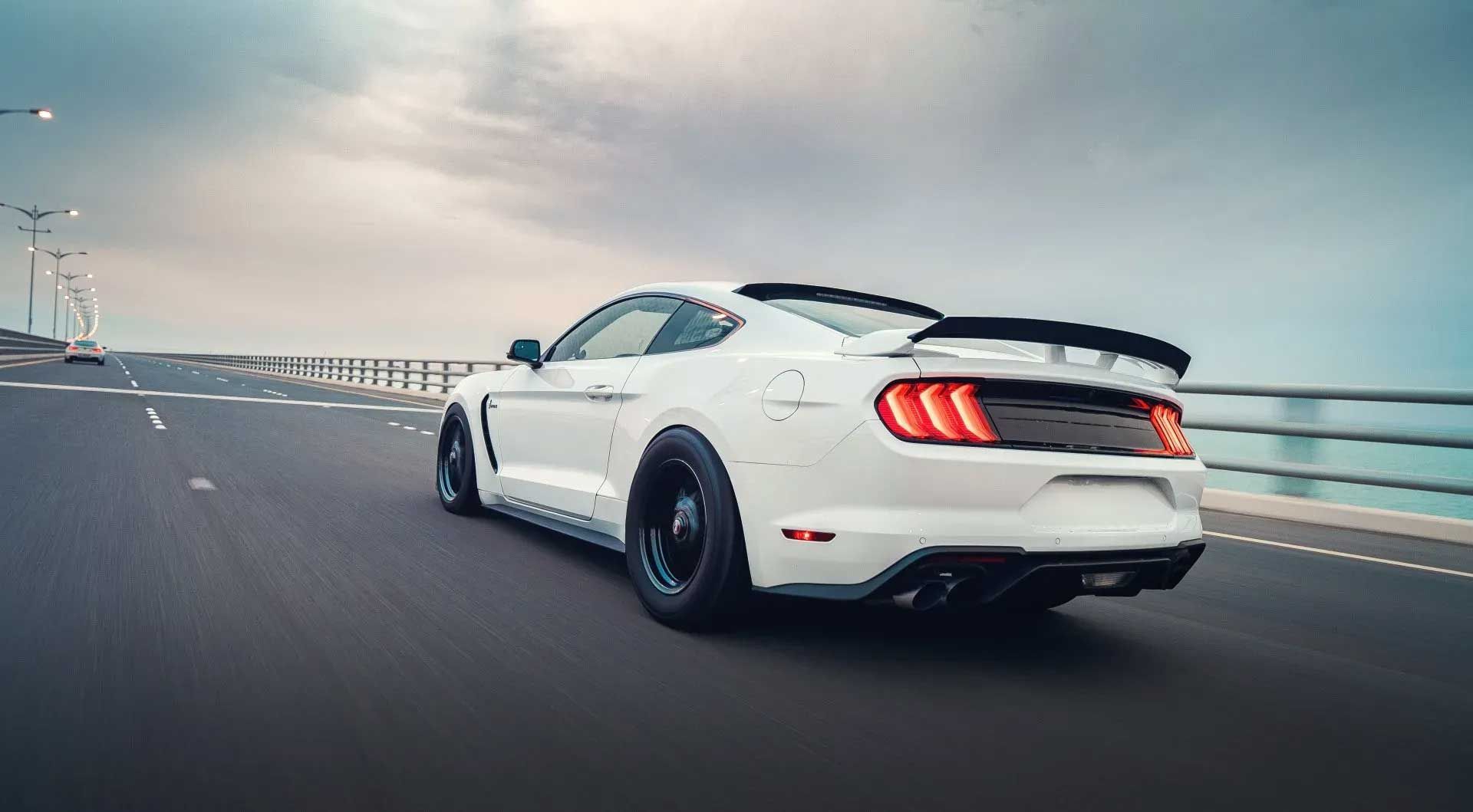 White Ford Mustang speeding on a bridge with a cloudy sky.