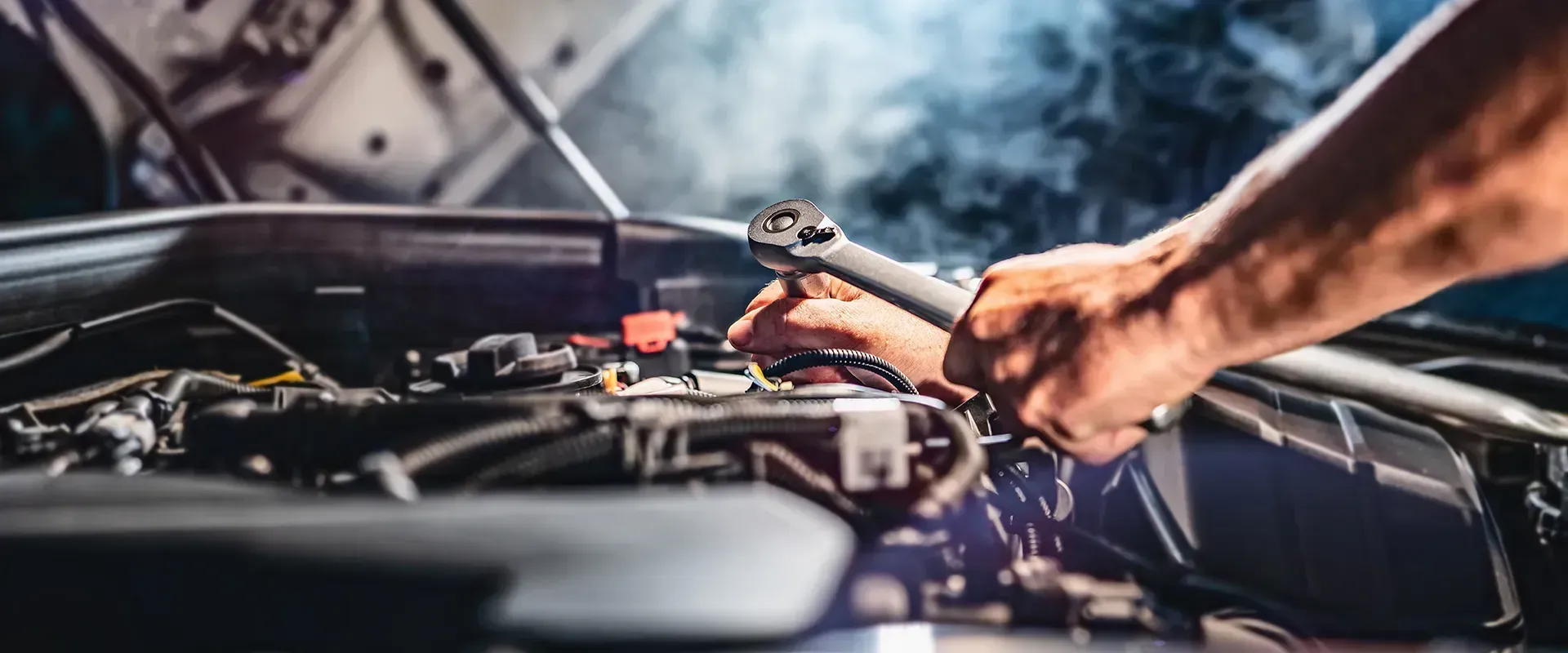 A person working on a car engine with a wrench. The hood is open, and smoke or mist is visible.