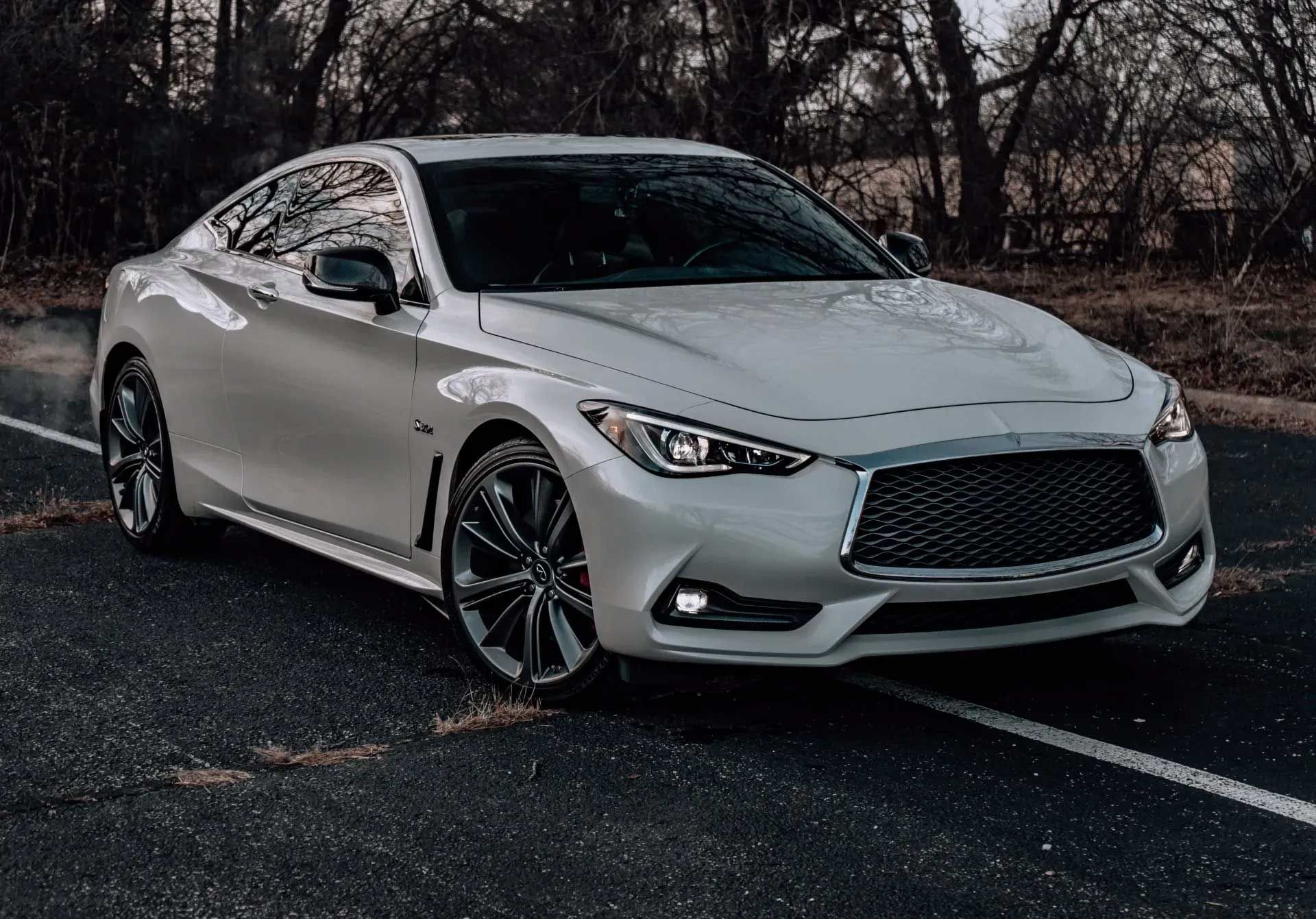 White Infiniti Q60 coupe on dark asphalt road, trees in background.