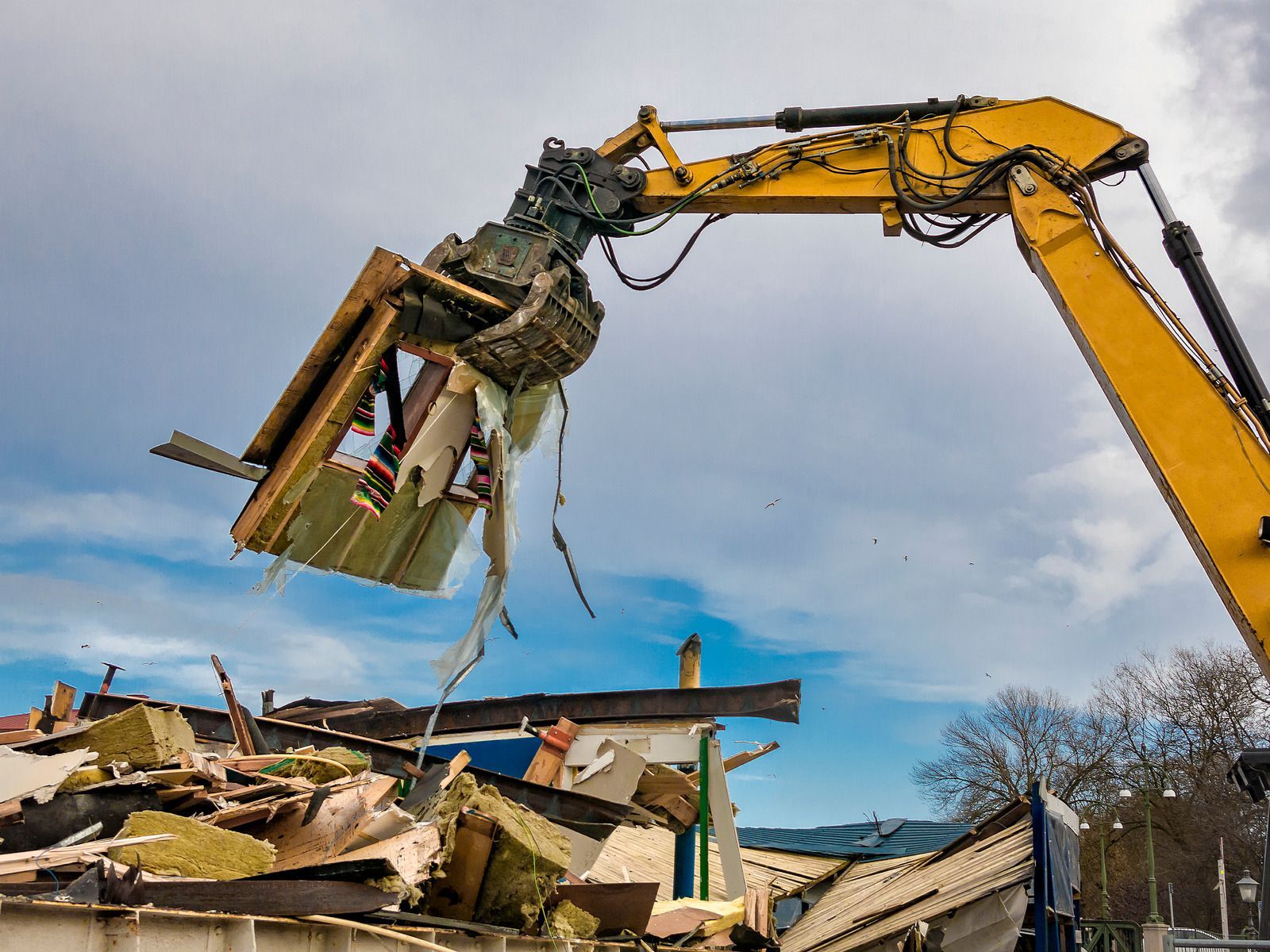 Yellow excavator demolishing a building, debris in foreground, cloudy sky.