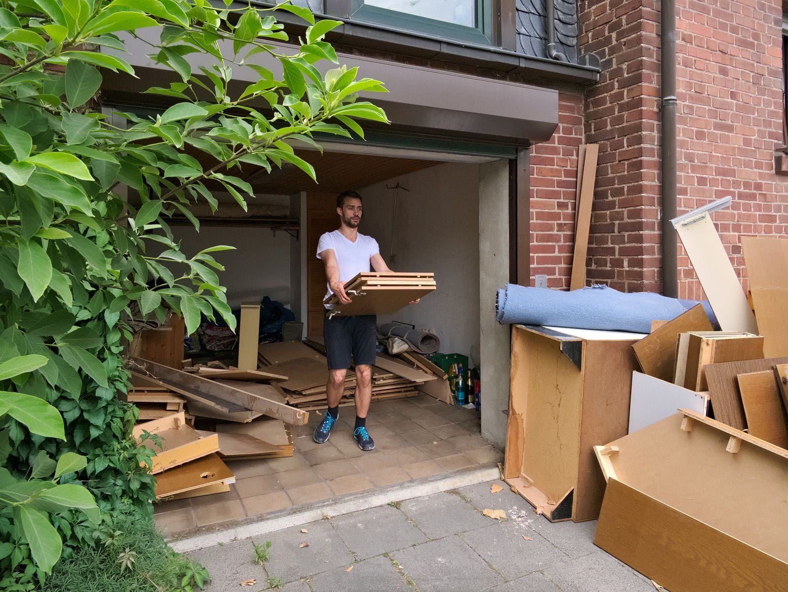 Man carrying wood out of a garage filled with debris; brick building, green foliage.