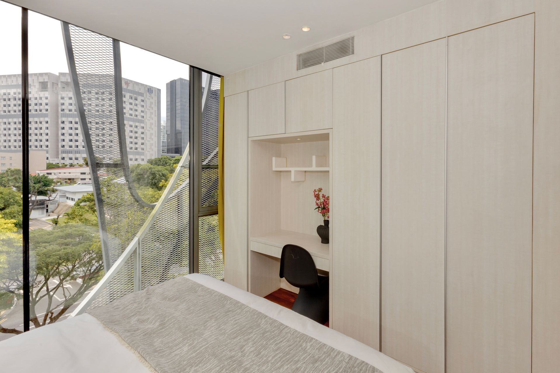 Bedroom with city view, built-in desk, and white storage cabinets. Black chair and red flowers visible.