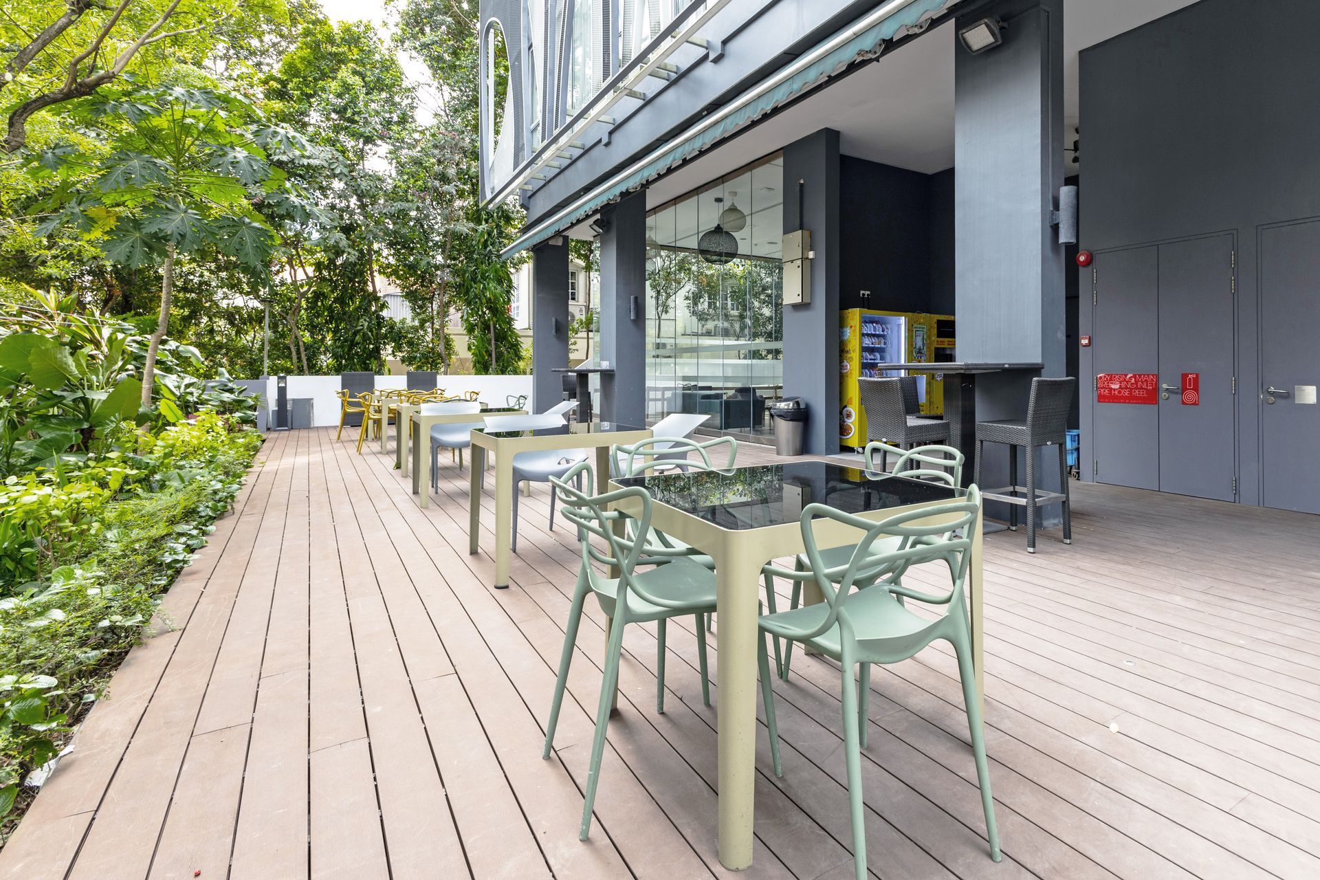 Outdoor cafe with green chairs, tables, and wooden deck, next to a building with trees.