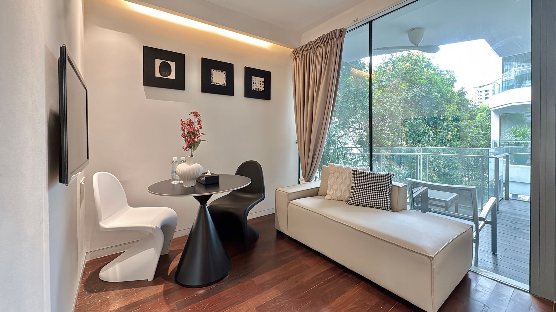 Modern living room with white walls, wood floors, and a balcony view. Features a small table, chairs, and a white chaise lounge.