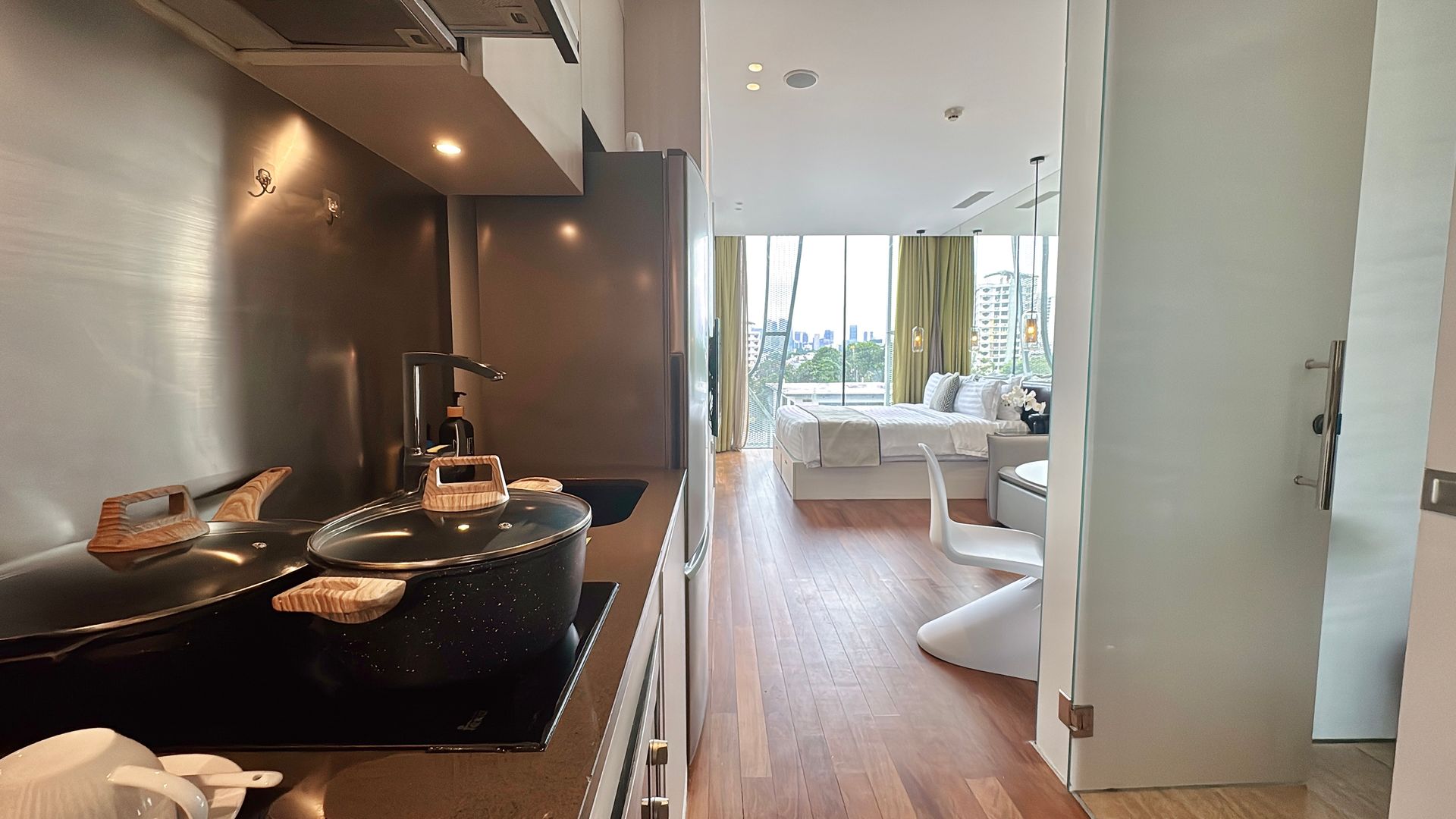 Kitchen view into modern studio apartment. Pot on stove, bed, white chairs, city view.