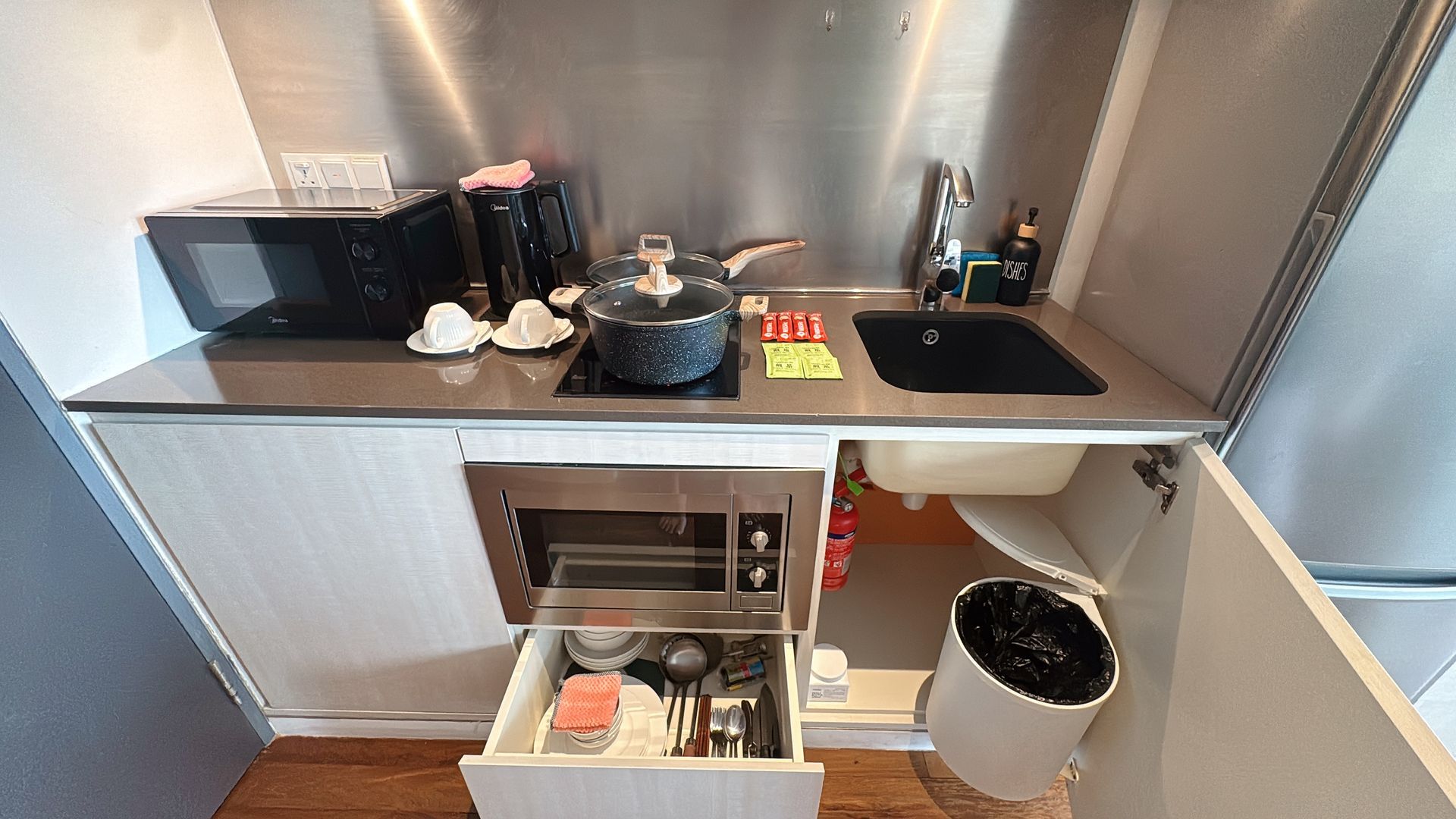 Small kitchen with stainless steel backsplash, microwave, sink, and open drawer with dishes.