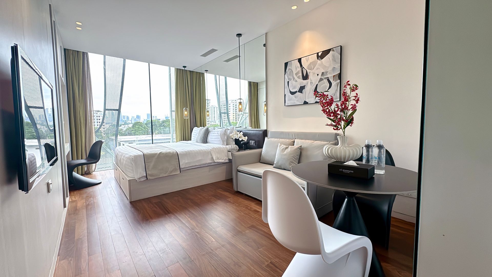 Modern studio apartment with large windows, bed, sofa, table, and decor.