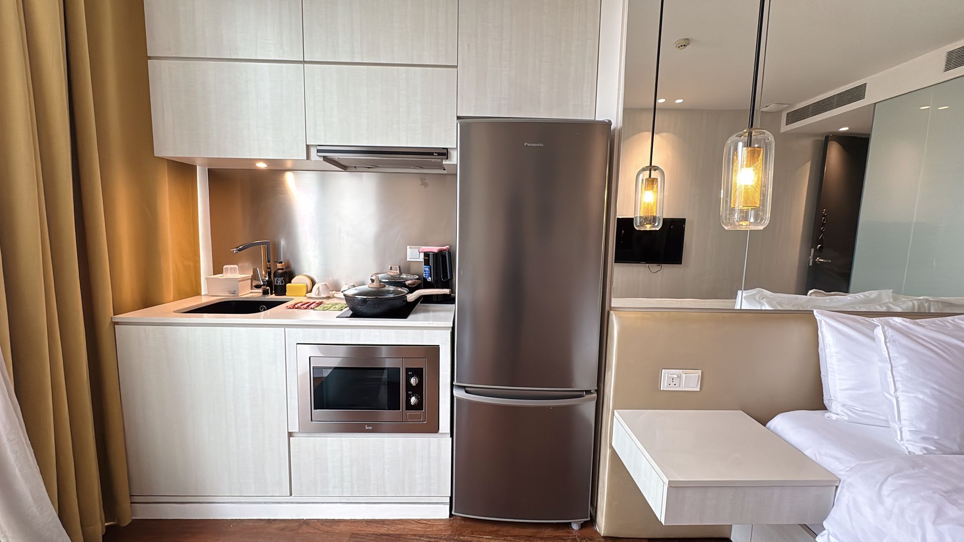 Small studio apartment with kitchenette, refrigerator, microwave, and bed. Beige and white color scheme.