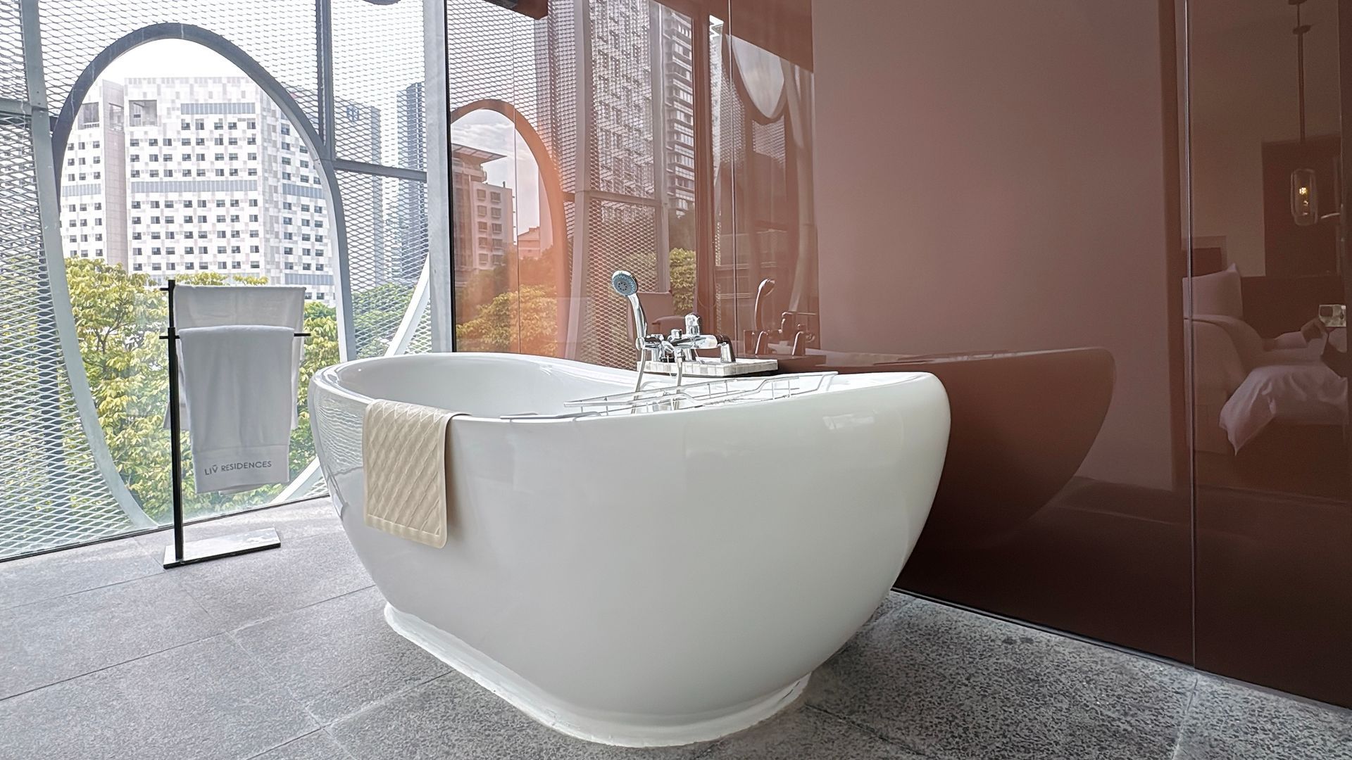 A white, oval bathtub with bubbles, in a modern bathroom overlooking a city through a large window.