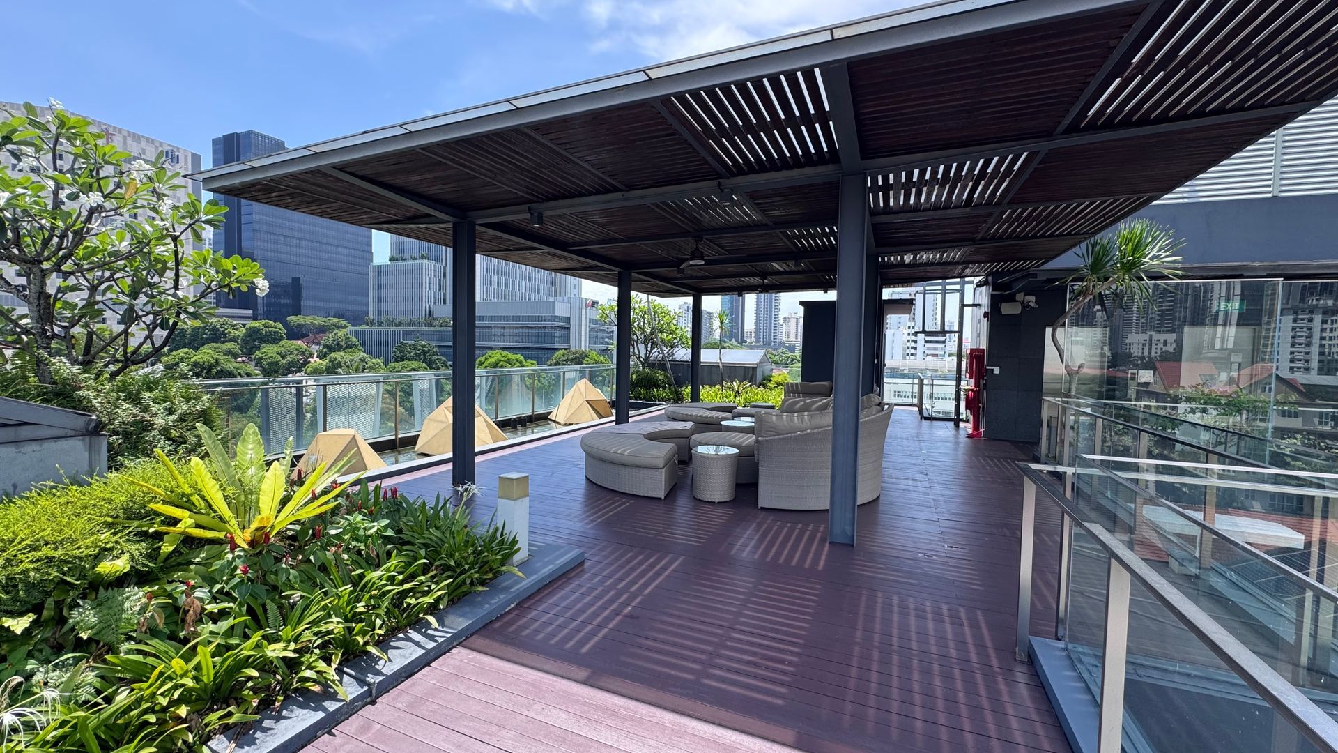 Rooftop patio with shaded seating area, surrounded by plants, with a city skyline view on a sunny day.