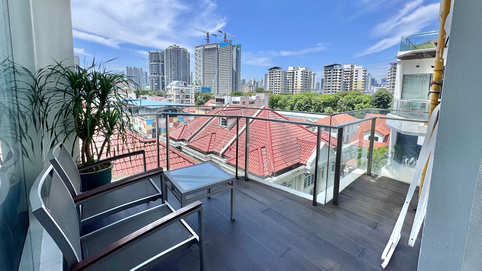 Balcony with two chairs, small table, potted plant, and cityscape view of red rooftops and modern buildings under a bright blue sky.