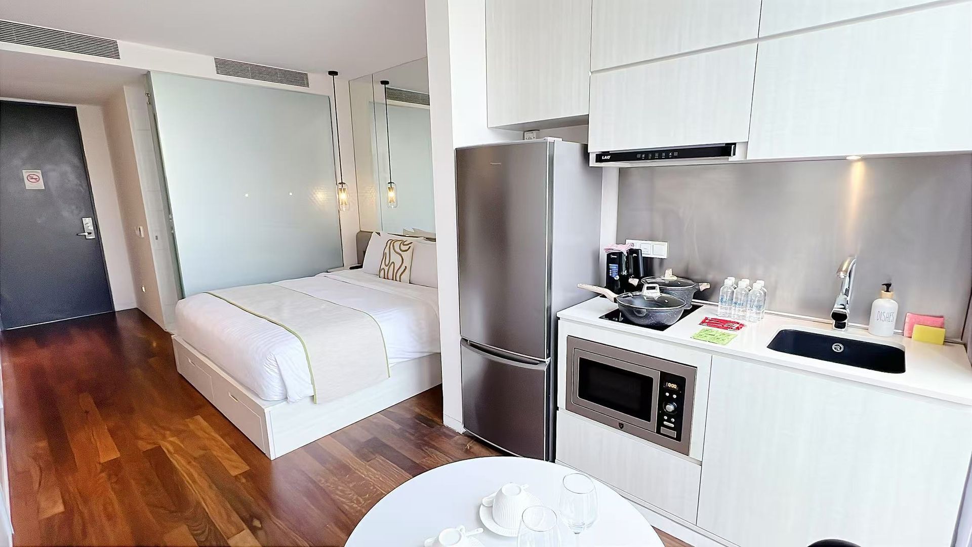 Studio apartment with kitchen, bed, and small dining table. White and wood tones throughout.