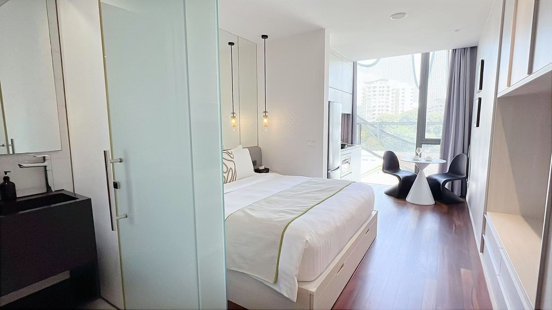 Modern bedroom with open bathroom and large window. Features bed, sink, and seating area.