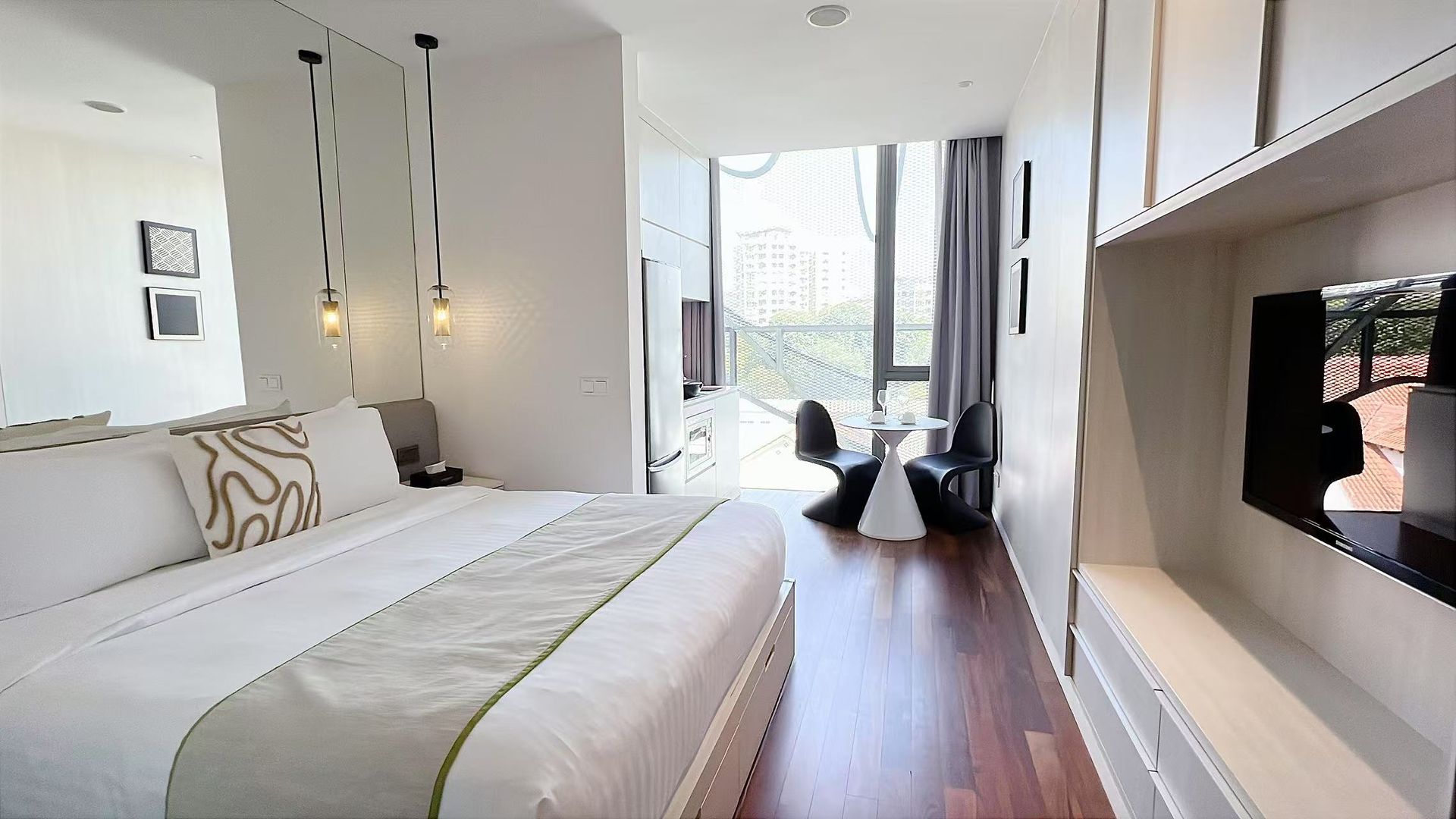 Modern hotel room with bed, TV, table, and large window; white, wood, and green accents.