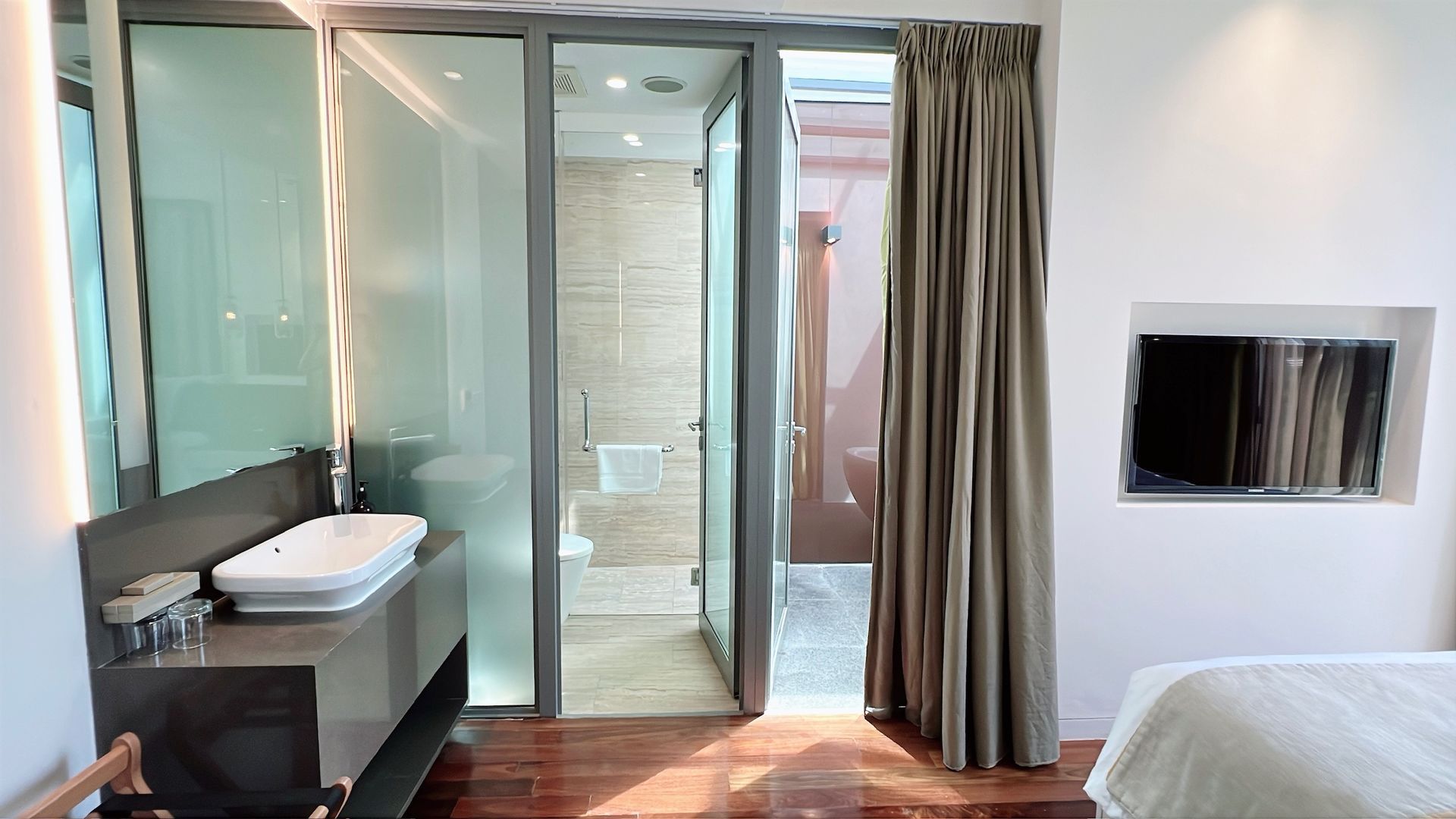Bathroom and bedroom combination. Sink, shower, curtain, and a wall-mounted TV in a hotel room.