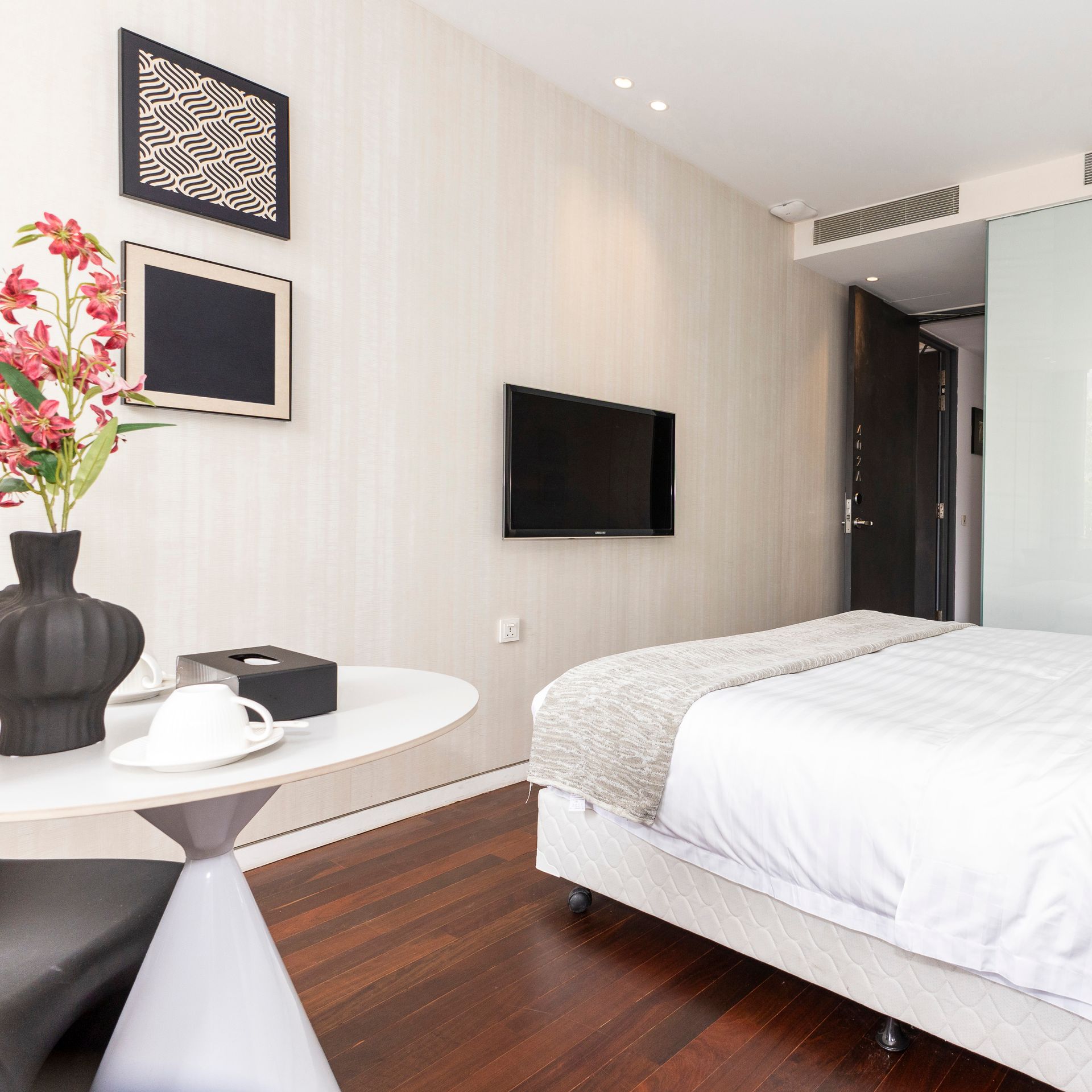 Modern hotel room with bed, TV, art, table with flowers, and a dark door.