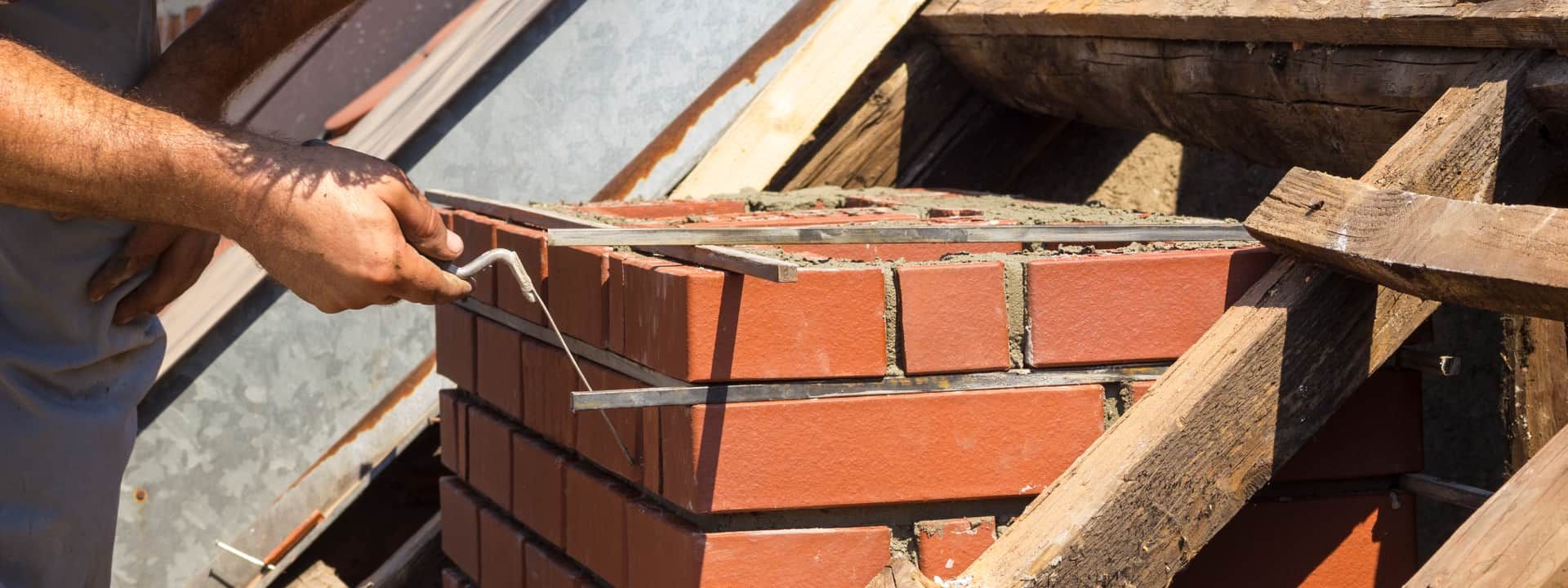 A person constructing a brick chimney on a roof. They are using a trowel-like tool.